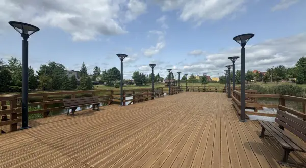 Drewniana promenada w Swarożynie z ławkami i latarniami, nad spokojną wodą, pod błękitnym niebem z chmurami.