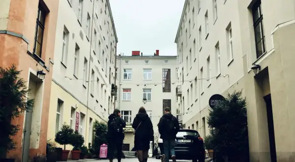Spacer po urokliwej uliczce w południe Polski. Trzy osoby idą w stronę kamienic, a w tle widać samochód i szyld kawiarni.