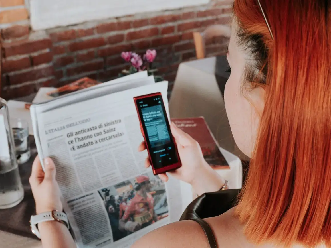 Kobieta z rudymi włosami używa telefonu do tłumaczenia tekstu z gazety. To przydatna wskazówka, jak poradzić sobie na lotnisku bez znajomości języka.