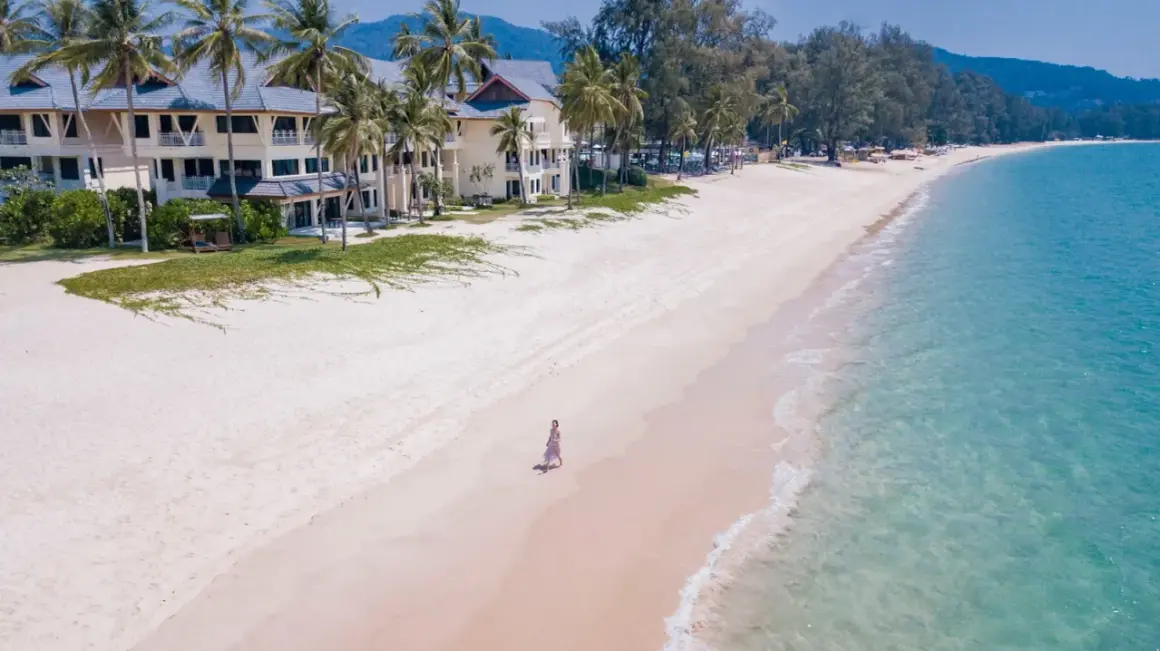 Kobieta spaceruje po piaszczystej plaży Bang Tao, otoczonej palmami i luksusowym hotelem.