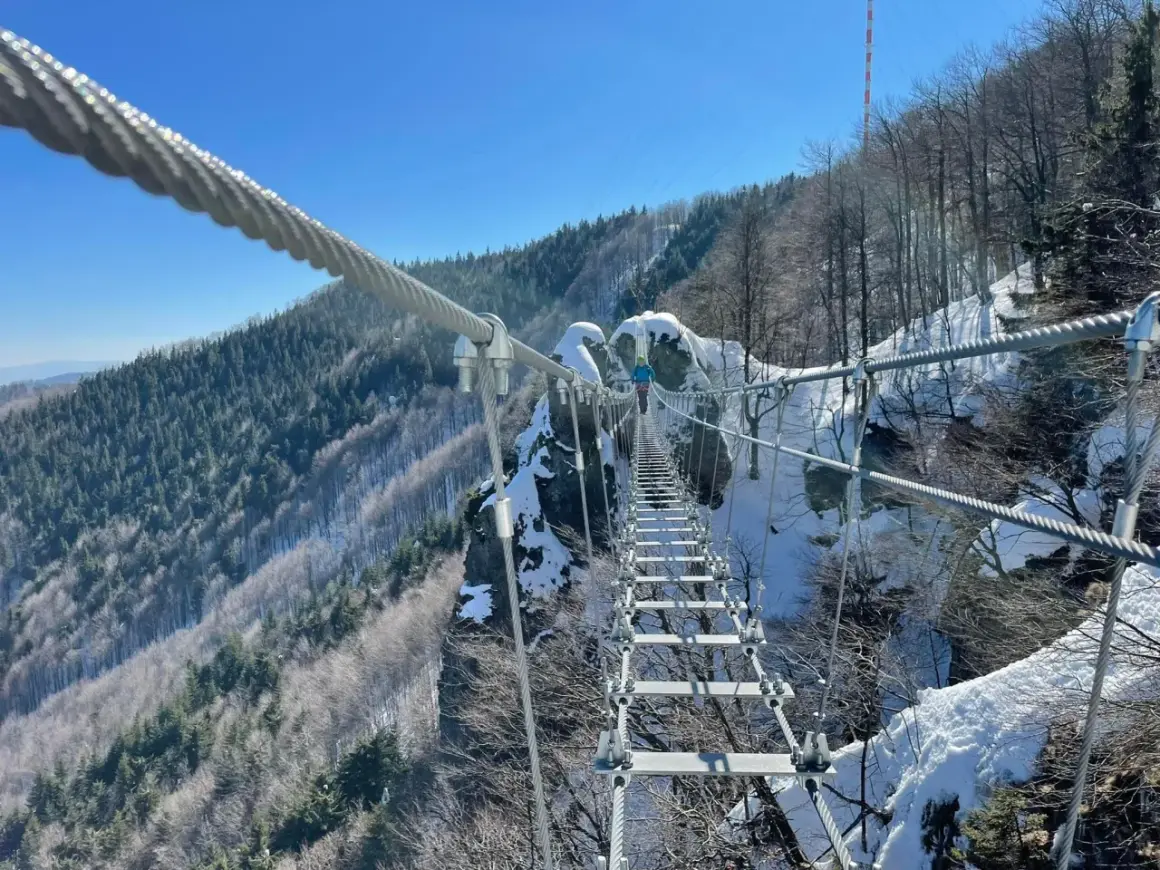 Osoba pokonuje most wiszący na via ferrata polska. Zimowy krajobraz górski, śnieg i gołe drzewa.