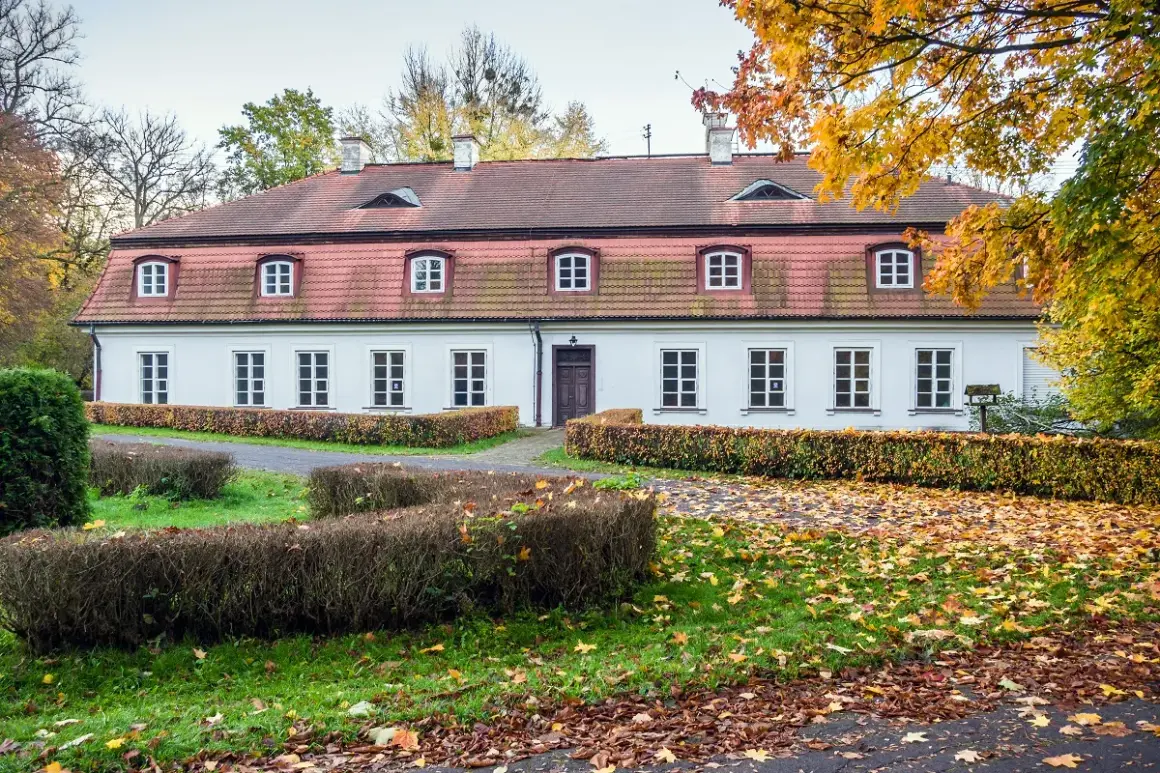 Biały dworek z czerwonym dachem, otoczony jesiennymi drzewami i krzewami. Wokół leżą opadłe liście, tworząc malowniczy krajobraz. Dawniej w takich miejscach znajdowały się obory.