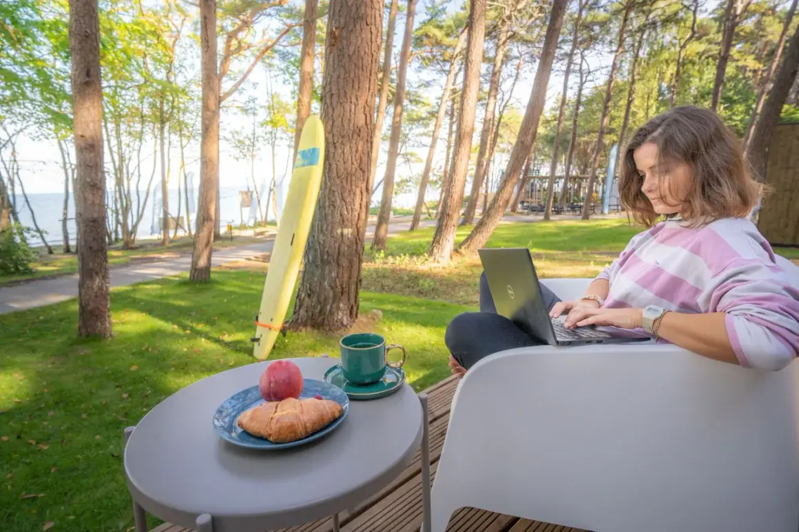 Kobieta pracuje na laptopie przy stoliku z kawą i croissantem. W tle widać morze i sosny, idealne miejsce dla domków z wyjściem na plażę.