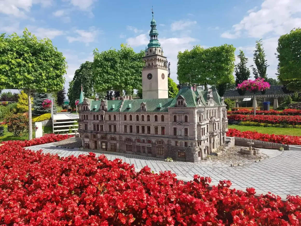 Miniatura ratusza z wieżą zegarową, otoczona czerwonymi kwiatami, przypomina o architekturze Kotliny Kłodzkiej.