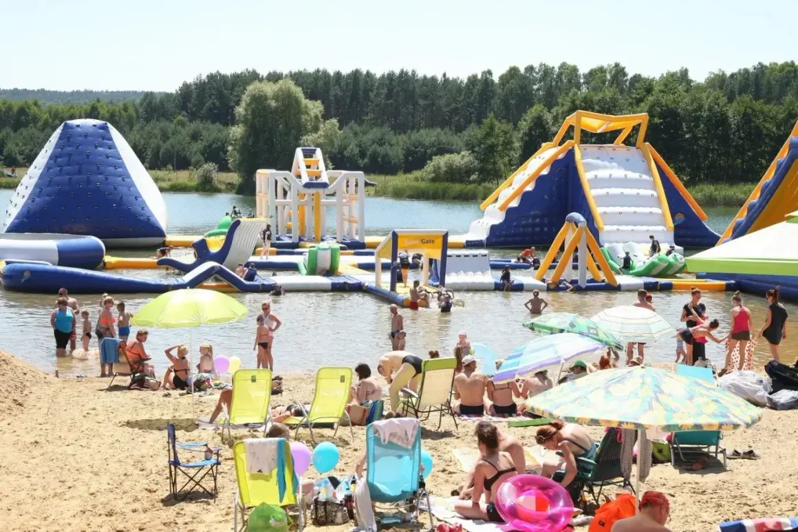 Rodzinny wypoczynek nad wodą na **bolmin zalew** z dmuchanymi atrakcjami wodnymi, zjeżdżalniami i placem zabaw. Słoneczny dzień, plaża pełna ludzi.
