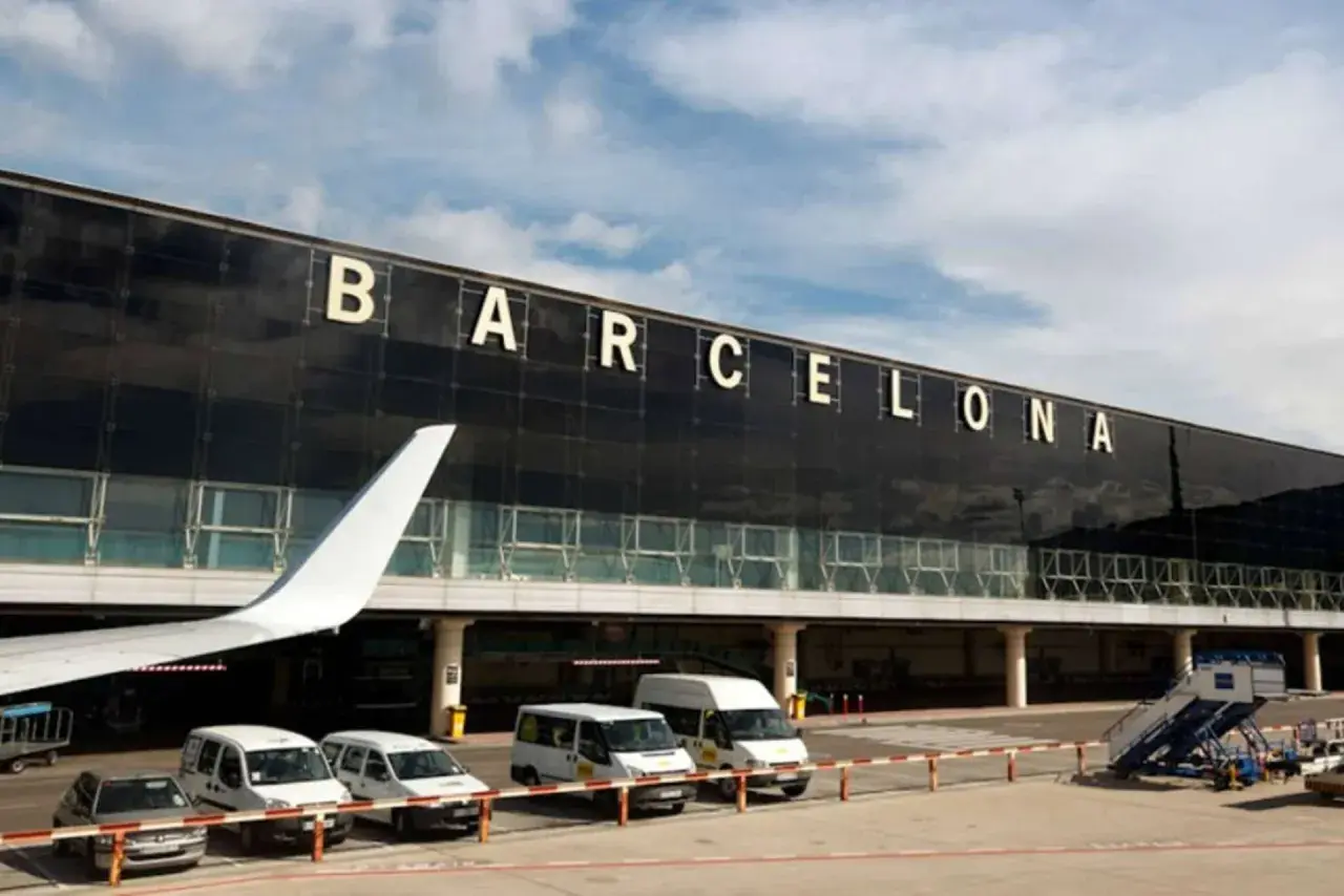 Duży napis BARCELONA nad nowoczesnym terminalem lotniska. Samolotowy skrzydło i busy czekają na pasażerów.