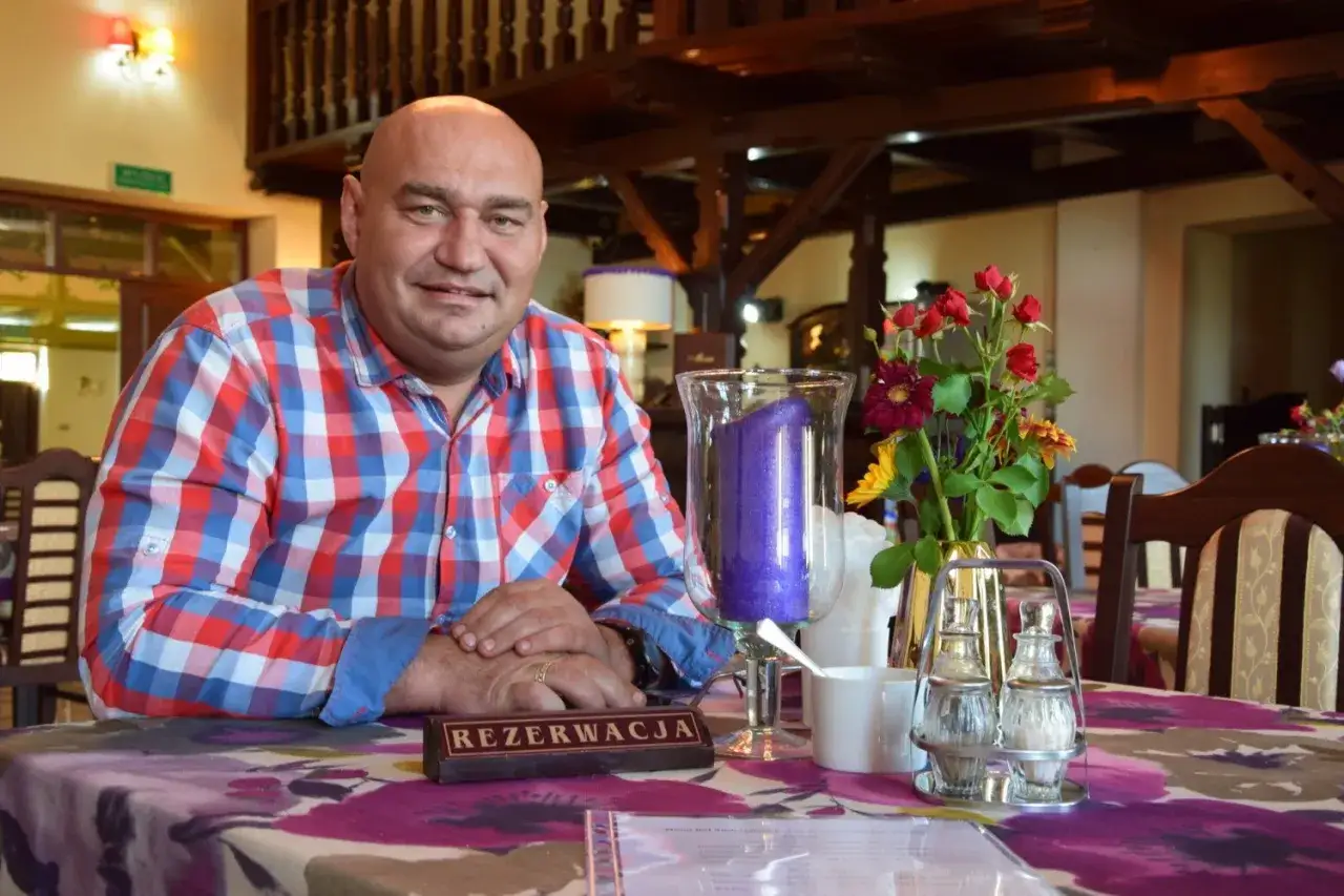Łysy mężczyzna w kratę siedzi przy stole w karczmie. Na stole wazon z kwiatami i świeca.