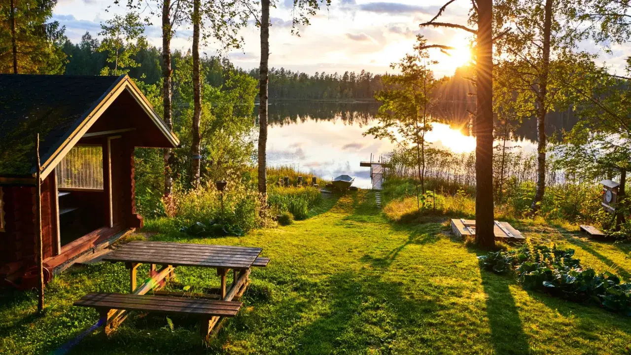 Sielankowy domek nad jeziorem w Finlandii. Słońce zachodzi, rzucając złote światło na trawę i wodę. Ceny noclegów tutaj są zaskakująco przystępne.
