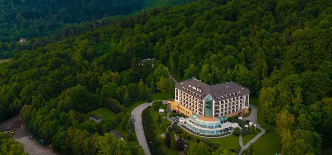 Uzdrowisko w Beskidzie Śląskim otoczone bujnym lasem. Nowoczesny budynek hotelowy z przeszklonym atrium wkomponowany w zielone wzgórza.