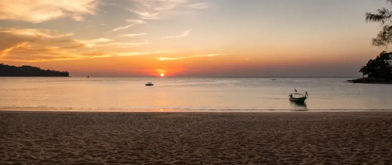 Zachód słońca nad plażą Bang Tao. Spokojne morze, łódź kołysze się na falach, a niebo mieni się odcieniami pomarańczy i różu.