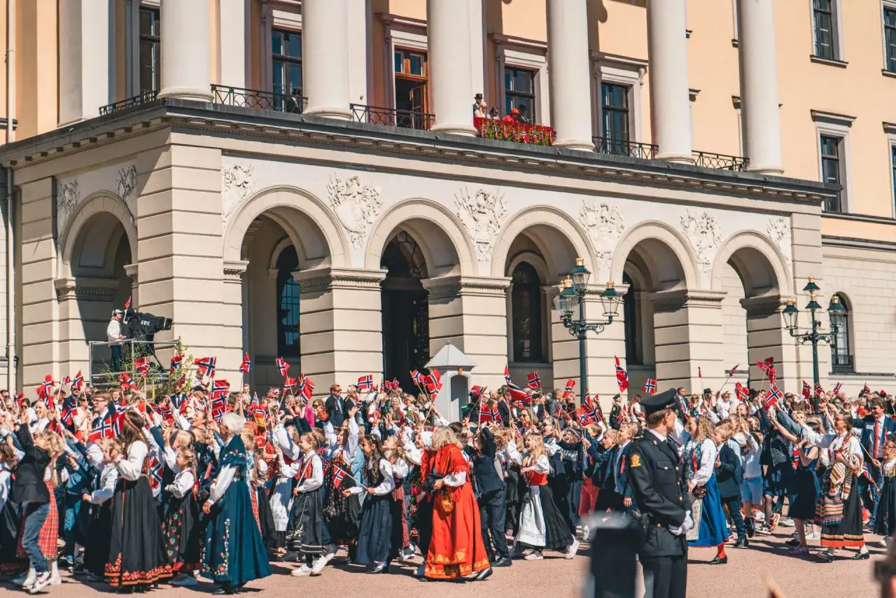 Tłum ludzi, w tym dzieci w strojach ludowych, świętuje na ulicy, machając norweskimi flagami. To radosne święta w Norwegii!