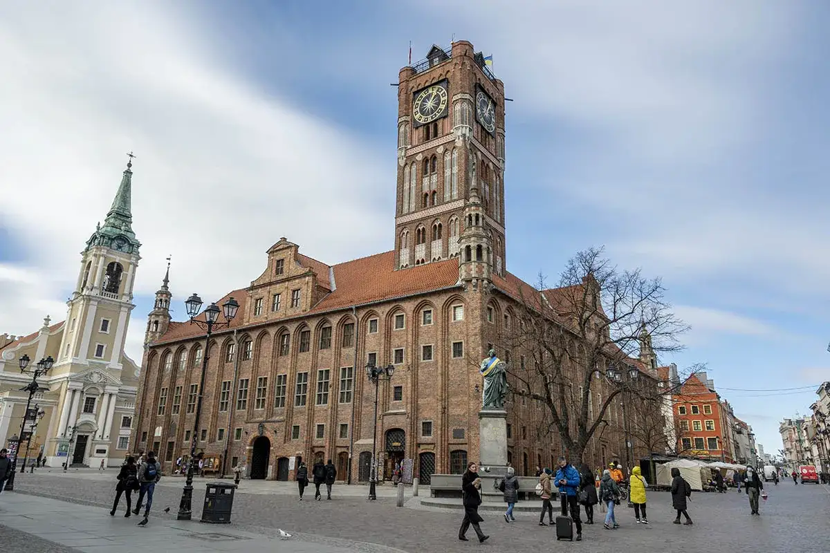 Rynek Staromiejski w Toruniu z Ratuszem i kościołem. Ludzie spacerują po placu, podziwiając zabytkową architekturę.