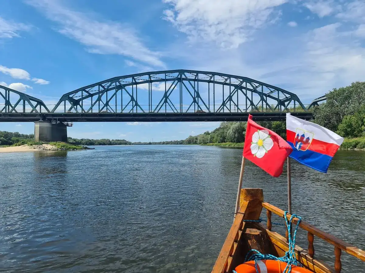 Rejs łodzią pod mostem, z widokiem na rzekę i dwa flagi. W tle zielony fordon.